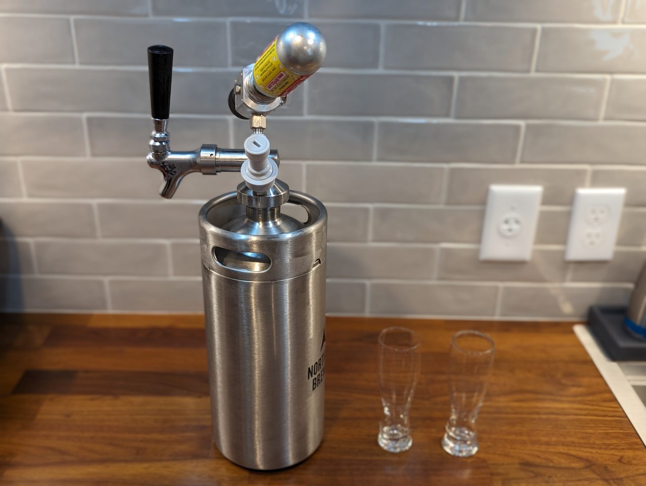 Photo of a one-gallon stainless steel keg with two small tasting glasses next to it