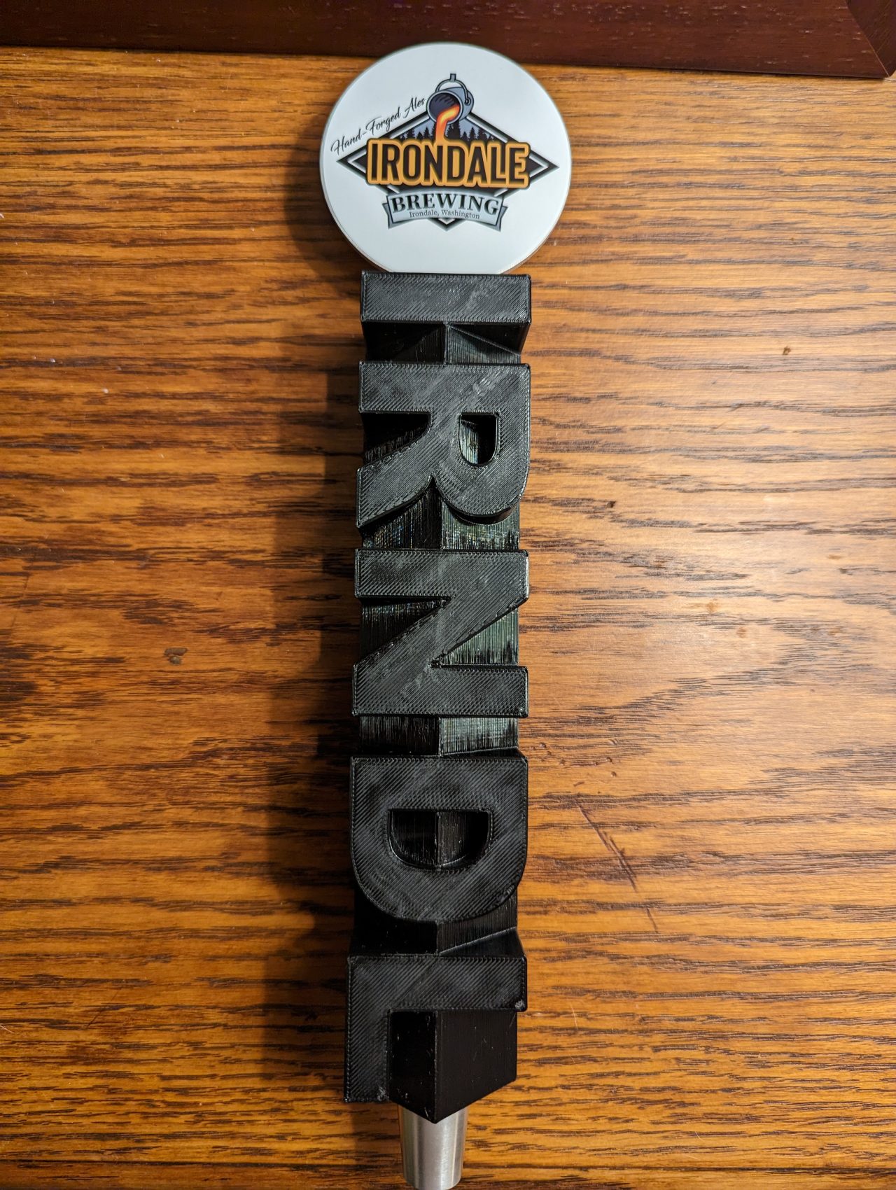 Photo of a tap handle from Irondale Brewing with the brand's logo at the top and the word "IRNDL" vertically spelled in large, black, 3D letters against a wooden background.