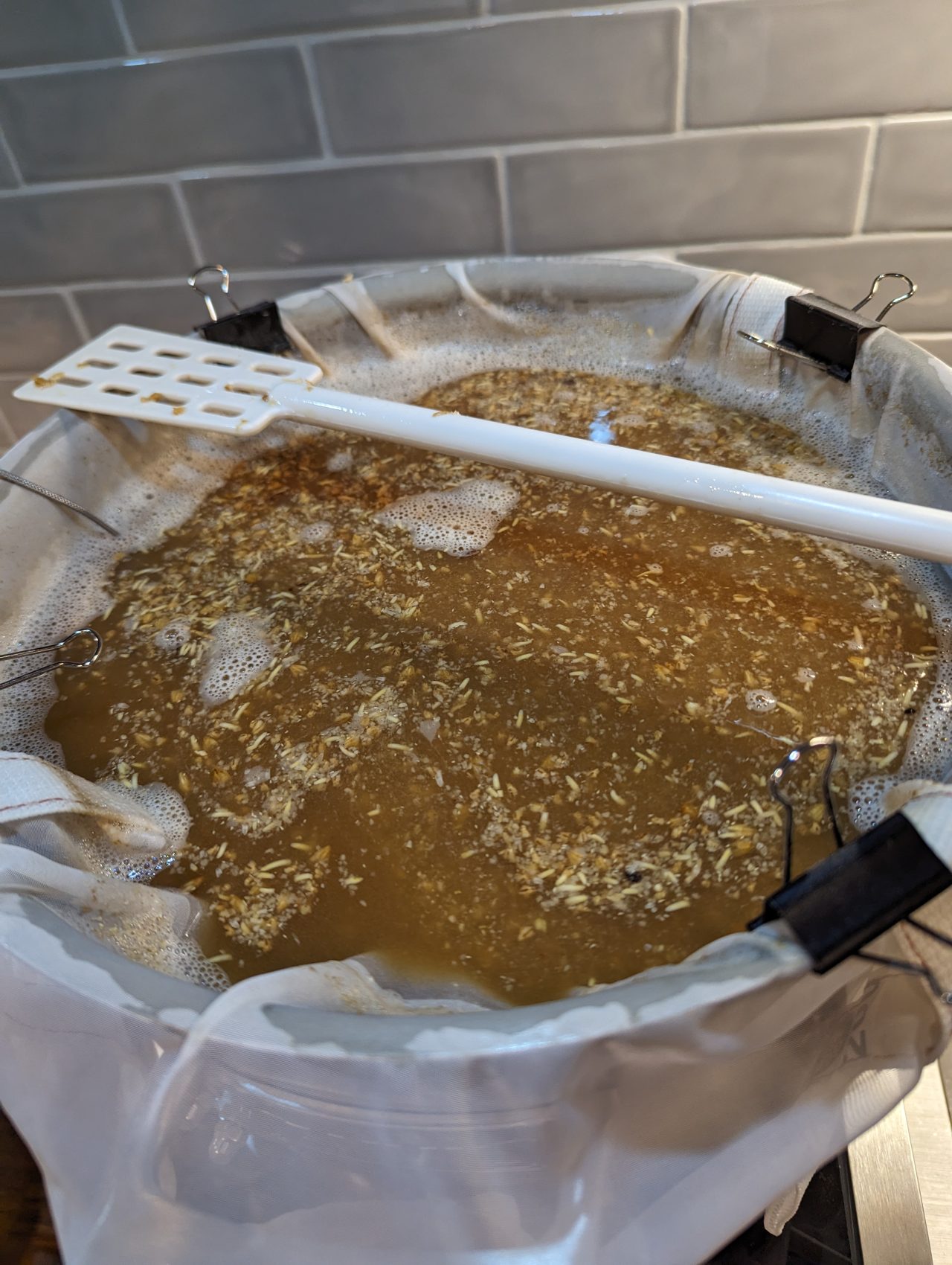 Photo of a 5-gallon brew pot filled with grains in a brew bag and a plastic mash paddle across the top